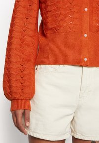 Pomarańczowy sweter z dzianiny o teksturowanym wzorze i ściągaczami przy mankietach. Posiada guziki przypominające perły. W połączeniu z białymi szortami dżinsowymi.