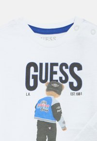 Λευκό βαμβακερό μπλουζάκι με μπλε λαιμόκοψη, που απεικονίζει μια αρκούδα με μπλε σακάκι και μαύρο παντελόνι, με το λογότυπο "GUESS" εμφανώς τοποθετημένο.