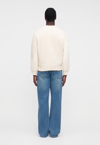 Άτομο που στέκεται, φοράει απλό κρεμ φούτερ και φαρδιά μπλε τζιν, κοιτάζοντας προς τα πίσω με φόντο ένα απλό ανοιχτόχρωμο φόντο.