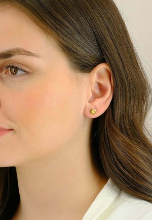 Orecchino a perno a forma di cuore in oro esposto sull'orecchio di un modello, con una finitura liscia e lucida e dimensioni ridotte, che sottolinea il design minimalista.