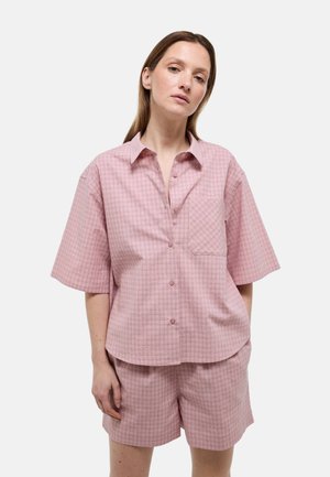 Femme portant une chemise ample à manches courtes boutonnée à carreaux rose avec un short assorti, debout devant un fond uni.