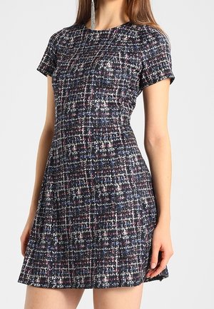 Femme portant une robe courte à manches courtes, ajustée, à carreaux noirs et blancs avec des touches de bleu et de rouge, posant devant un fond uni.