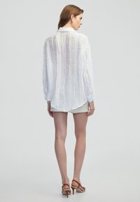 Blouse blanche texturée avec une coupe décontractée, dotée d'un ourlet asymétrique et de manches longues, assortie à un short beige et des sandales à talons couleur tan.