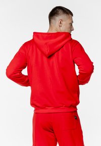 Czerwona bluza z kapturem o fakturowanej powierzchni. Posiada kieszeń kangurzą i ściągacze na rękawach, noszona z pasującymi czerwonymi spodniami.