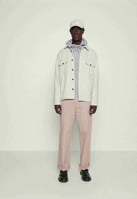 Giacca bianca con bottoni sopra a una felpa con cappuccio grigio chiaro, abbinata a pantaloni rosa chiaro e scarpe nere. Il modello indossa un cappellino bianco.