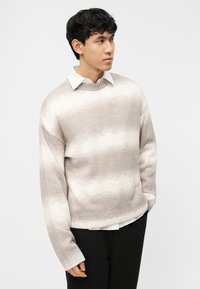 Jeune homme aux cheveux noirs portant un pull beige à rayures blanches sur une chemise blanche à col, associé à un pantalon noir, debout devant un fond blanc.