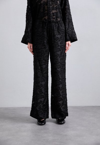 Îmbrăcăminte neagră transparentă, cu texte florale intricate, pantaloni larghi și mâneci lungi cu o croială lejeră. Pantofi negri simpli completează look-ul.