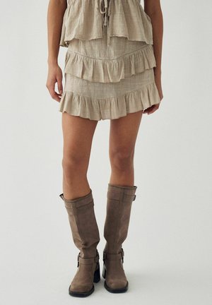 Kobieta ubrana w beżową, warstwową mini spódniczkę z falbanami oraz taupe wysokie kozaki na obcasie, stojąca na tle jasnego, jednolitego tła.