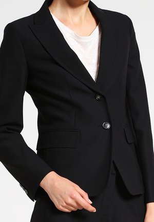 Kobieta ubrana w dopasowaną czarną marynarkę na dwa guziki z klapami kieszeni na białej koszuli, z ręką spoczywającą niedaleko talii.