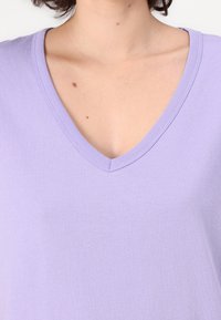 Lila V-nyakú póló, puha pamutból készült. Simaság jellemzi, testhezálló fazonnal, letisztult dekoltázzsal és finom varrási részletekkel.
