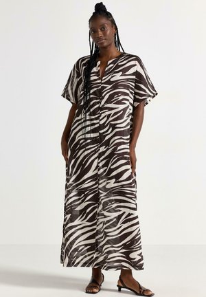 Femme portant une robe longue ample imprimée zèbre noir et blanc avec manches courtes et des sandales noires à petit talon, debout devant un fond uni.
