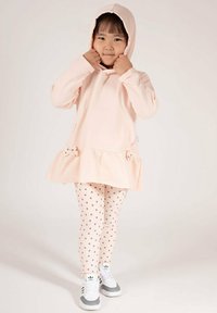 Jasnoróżowa bluza z kapturem z rozkloszowanym dołem i ozdobami w formie kokardek, zestawiona z kremowymi legginsami w kropki oraz szaro-białymi sneakersami. Codzienny strój dziecięcy.