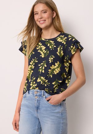 Femme souriante aux longs cheveux blonds porte un chemisier à manches courtes bleu marine à motif floral et un jean taille haute bleu clair, debout avec une main dans la poche.