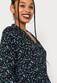 Vestido preto com flores pequenas multicoloridas, decote em V e mangas longas. O tecido tem uma aparência texturizada. Grandes brincos de argola.