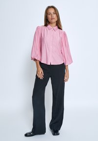 Camicia rosa a maniche puff con texture morbida e colletto, abbinata a pantaloni neri a gamba larga e scarpe nere lucide.