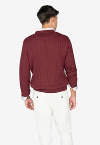 Maglione bordeaux a maglia grossa con colletto rotondo, orlo a costine e maniche lunghe, indossato sopra pantaloni bianchi, visto da dietro.