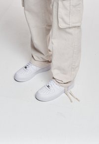 Personne portant un pantalon cargo beige en velours côtelé et des baskets blanches à lacets debout sur un sol blanc.