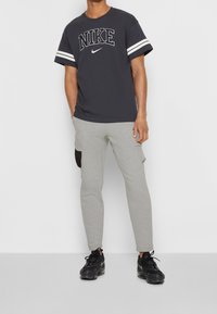 Černé tričko Nike s bílými pruhovanými rukávy a logem, doplněné šedými tepláky s černou boční kapsou a černými sportovními botami.