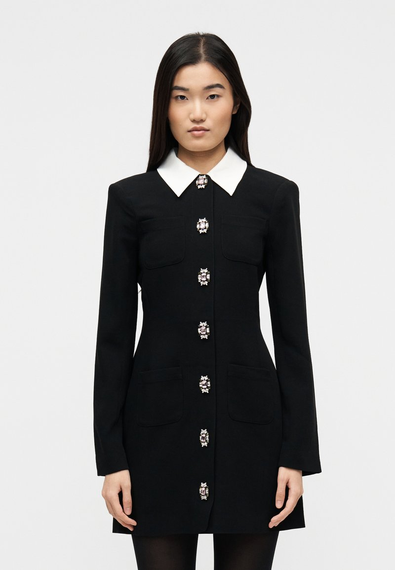 Γυναίκα που φοράει μαύρο φόρεμα με μακριά μανίκια, λευκό γιακά και περίτεχνα στολισμένα κουμπιά με πολύτιμους λίθους κατά μήκος του μπροστινού μέρους, στέκεται μπροστά σε λευκό φόντο.