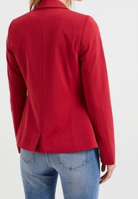 Röd skräddarsydd kavaj med slagkrage, enkel ryggslits och strukturerade axlar, i kombination med blå jeans. Slät tygtextur.