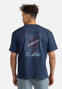 Námořnická modrá bavlněná tričko s retro-style grafikou zobrazující globe, mřížkový vzor a siluetu hor, doplněné červenými a akvamarínovými akcenty.