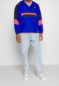 Modrá lehká bunda s černými a růžovými detaily, kapucí, částečným zipem vpředu a předním kapsou. Nošena přes růžové tričko a světlé džíny.