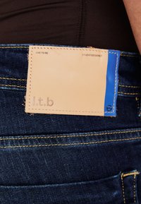 Modré riflové jeans s námornými švami a svetlým koženým štítkom na zadnej kapse, ktorý obsahuje embosovaný text a modrú akcentačnú časť.
