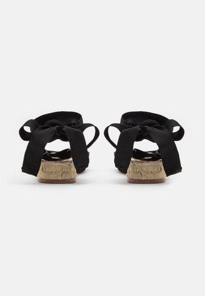 Μαύρες εσπαντρίγες με πλεκτή σόλα από γιούτα. Διαθέτουν φαρδείς μαύρους υφασμάτινους ιμάντες δεμένους σε φιόγκο στο πίσω μέρος. Υφή φινιρίσματος.