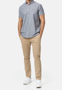 Camisa de manga corta, abotonada, en tela azul claro con una textura sutil, combinada con pantalones chinos beige de corte slim y zapatillas blancas.