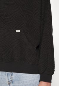 Pull-over noir en tissu texturé présentant un motif géométrique. Comprend une poche avant et des poignets et ourlet côtelés.