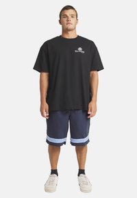 Czarny bawełniany t-shirt z okrągłym dekoltem, luźny krój. Granatowe szorty z niebieskimi paskami na końcu. Białe sneakersy z teksturowaną podeszwą. Model stoi w pozycji wyprostowanej.