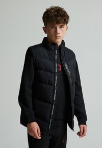 Chaqueta acolchada negra con cuello alto, cierre de cremallera y detalles de logo. La tela es suave con una sección acolchada texturizada en la parte delantera.