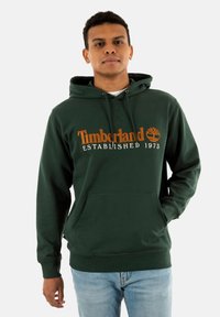 Sudadera verde con cordón ajustable, bolsillo canguro frontal y texto "Timberland Established 1973" en naranja. Tejido de mezcla de algodón.
