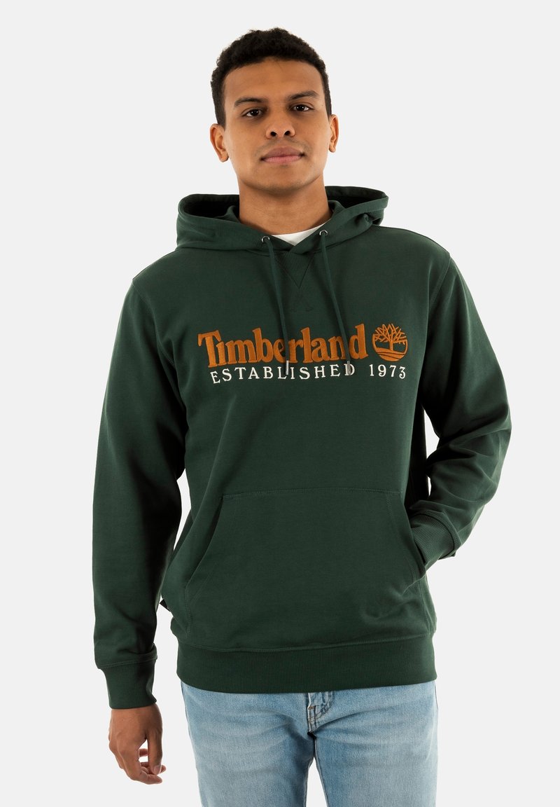 Sudadera verde con cordón ajustable, bolsillo canguro frontal y texto "Timberland Established 1973" en naranja. Tejido de mezcla de algodón.