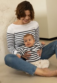 Gestreepte longsleeve shirts in marineblauw en wit, gedragen door een volwassene en een baby. De top van de volwassene heeft een gouden accent. Casual denim en een zandachtige achtergrond.