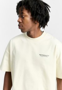 T-shirt kratkih rukava boje krem, izrađen od pamuka s opuštenim krojem. Sadrži tekst "RETERNITY CREATIVE DEPARTMENT" u crnom otisku.