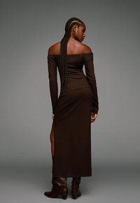 Rochie lungă, cu umerii goi, de culoare maro, cu o siluetă ajustată, crăpătură laterală și textură fină, asortată cu cizme maro până la genunchi.