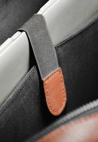 Sangle en tissu noir avec un accent en cuir brun tenant un ordinateur portable argent. Fermeture à zipper visible en bas, contrastant avec le matériau textile.