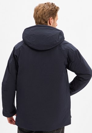 Giacca a vento blu navy con finitura liscia e opaca, caratterizzata da una vestibilità rilassata e un design semplice. Nessun logo o motivo visibile.