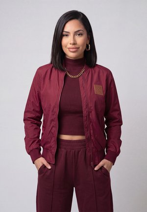 Woman with black hair wears a maroon jacket, cropped turtleneck, and pants, standing with hands in pockets against a plain background.