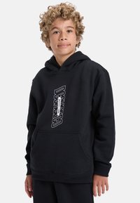 Čierna mikina s kapucňou, ktorá má predné klokanie vrecko a biely vertikálny potlač s logom. Mäkčený bavlnený materiál s textúrovaným povrchom.