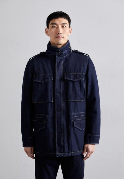 Giacca in denim blu scuro con colletto alto, due tasche sul petto e due tasche inferiori, con cuciture gialle a contrasto e accentuazioni sulle spalle.