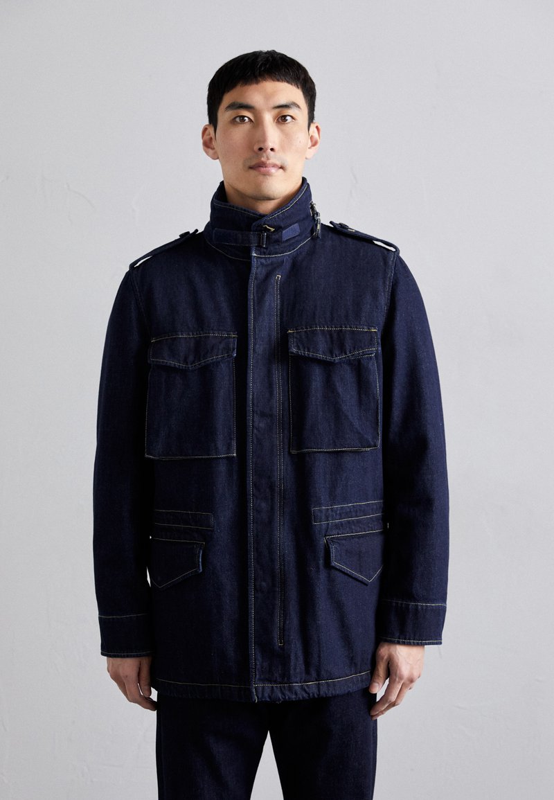 Dark blue denim jacket featuring a high collar, two chest pockets, and two lower pockets, with contrasting yellow stitching and shoulder accents.