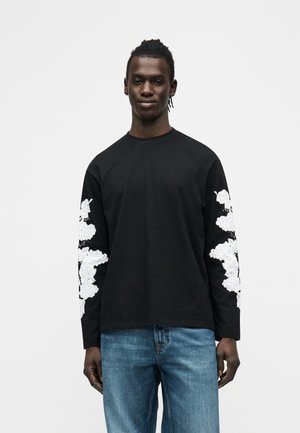 Junger Mann trägt ein schwarzes Langarmshirt mit weißen Blumenmustern auf den Ärmeln und blaue Jeans, steht vor einem schlichten weißen Hintergrund.