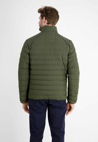 Olivgrüne Pufferjacke mit horizontal gestepptem Design, hohem Kragen und dezenten Schulternähten. Dazu werden marineblaue Hosen getragen.