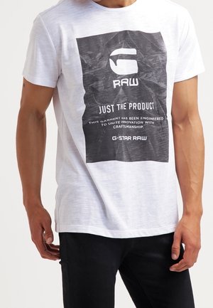Personne portant un t-shirt blanc avec une grande impression rectangulaire noire affichant le logo "G RAW" et un texte sur le savoir-faire, associé à un pantalon noir.