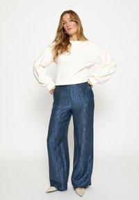 Witte katoenen trui met lange opgeblazen mouwen, gecombineerd met wijde metalen blauwe broek met een gestructureerde afwerking. Puntige beige schoenen.