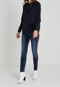 Marinblå sweatshirt med V-formad halsringning prydd med nitar och 'RR'-tryck, kombinerad med mörkblå jeans med röda randiga sömmar i sidan och vita ankelboots.
