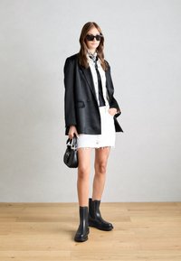 Blazer negro oversize, camisa blanca de botones debajo de un top negro sin mangas, falda de mezclilla blanca deshilachada, botas negras hasta el tobillo y bolso negro pequeño.