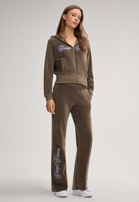 Bruin velours trainingspak met een rits-capuchon, zijzakken en geborduurde tekst. Losse broek met brede pijpen en logo-detail op het been.